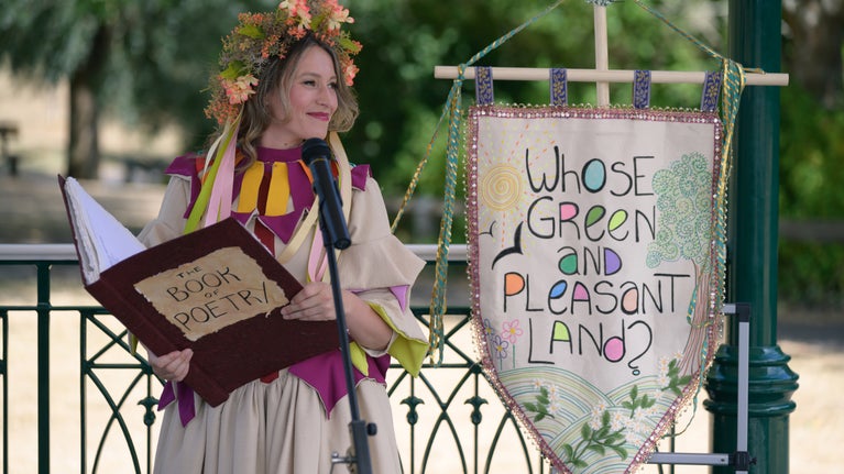 Woman in a cream and red dress and a head garland, reading from a large red poetry book, standing next to a cloth banner embroidered with the words: Whose green and pleasant land?